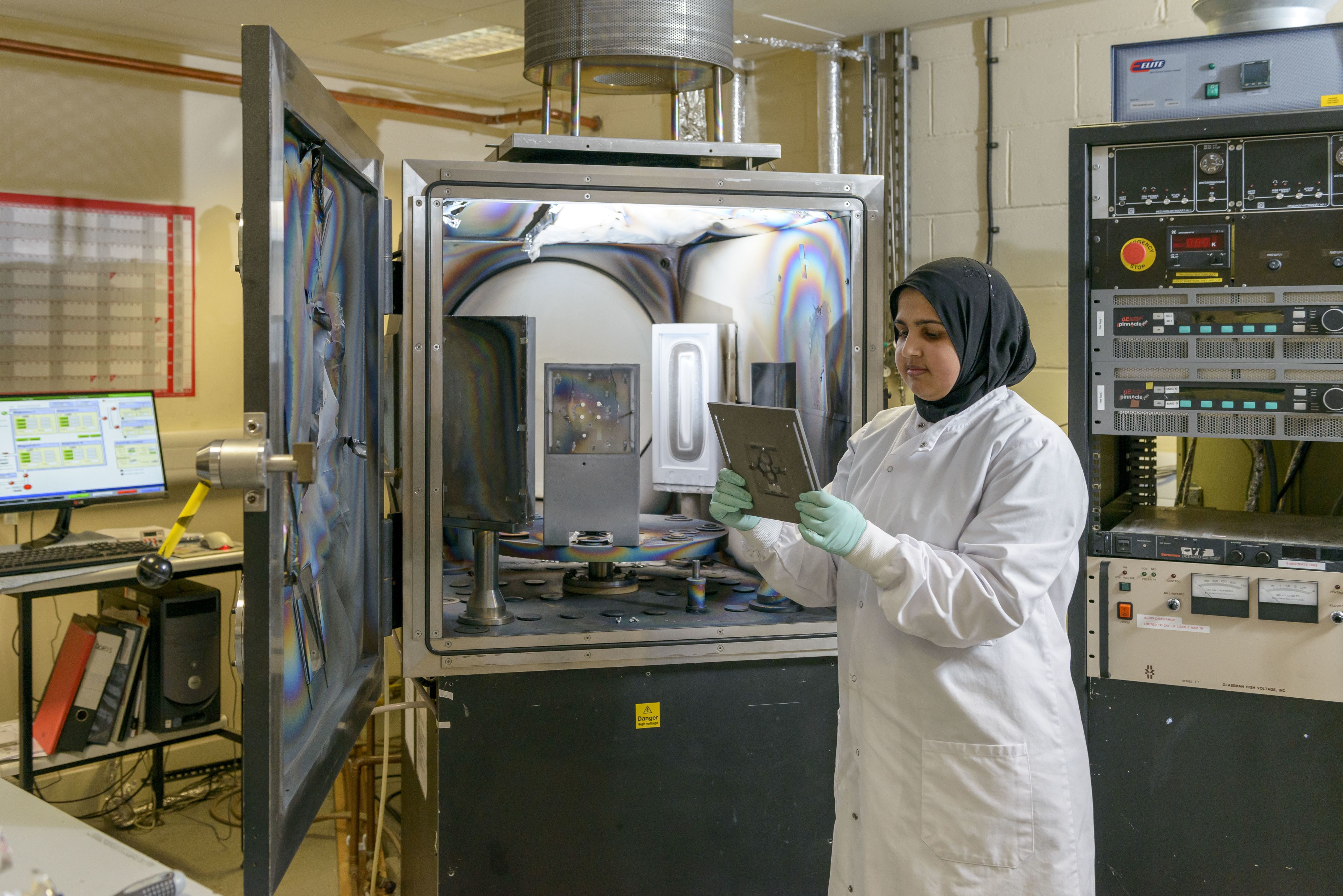 Man in Cranfield University blue lab coat with purple clove holding sample in front of dual source barrel physical vapour deposition facility