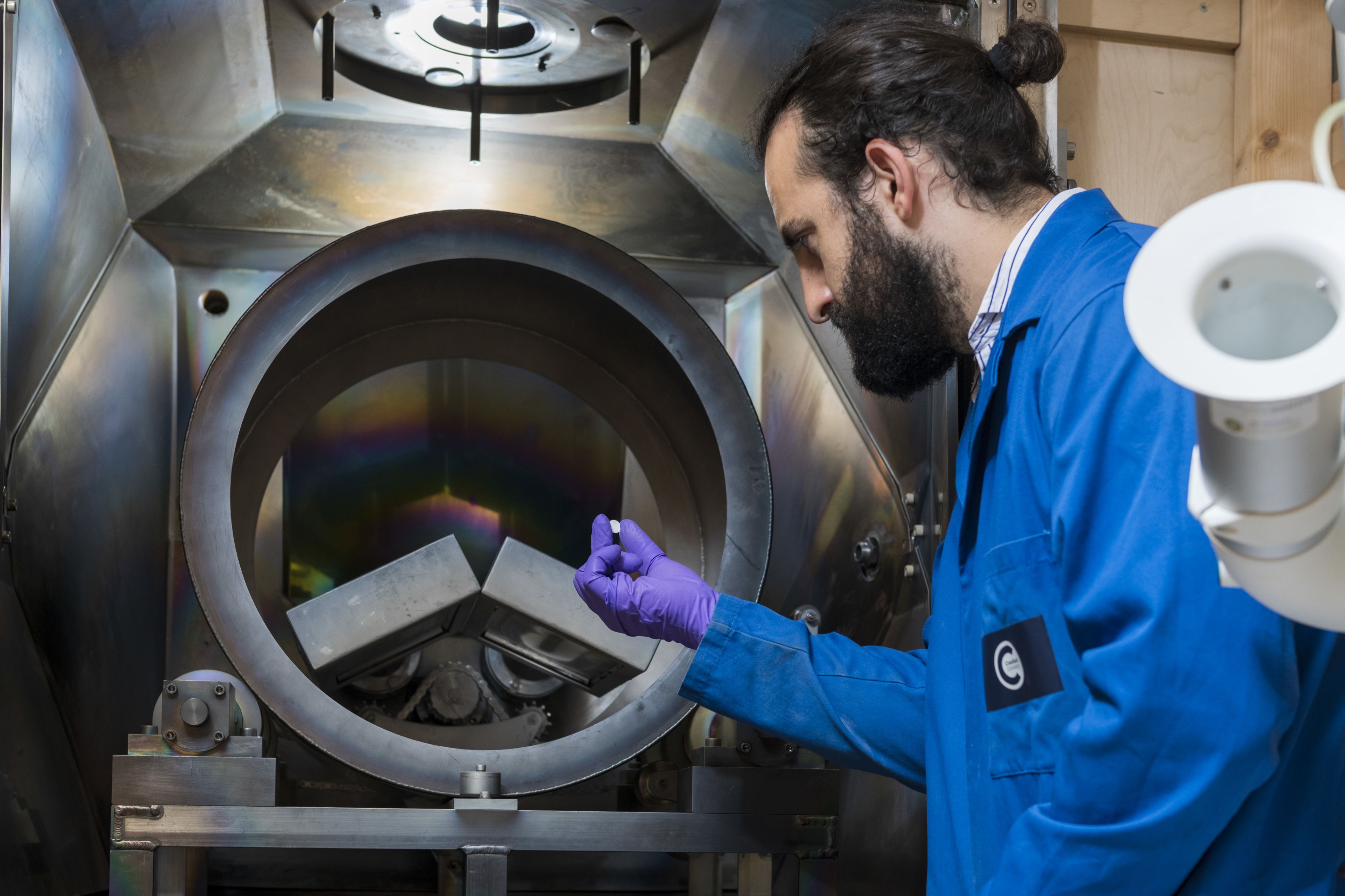 Man in Cranfield University blue lab coat with purple clove holding sample in front of dual source barrel physical vapour deposition facility